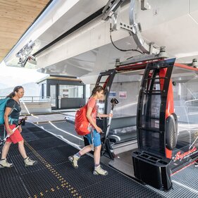 Zwei Frauen mit Wanderausrüstung steigen in eine Gondel ein